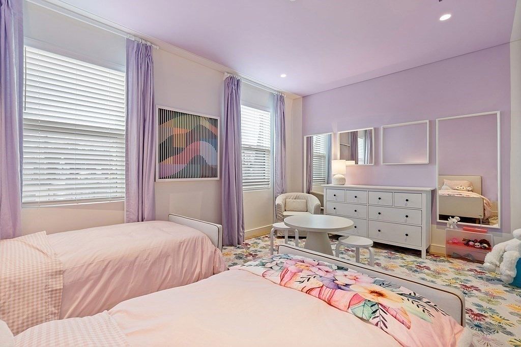 This is a charming children's bedroom featuring two twin beds with pink bedding and floral accents. The room is painted in a soft lavender hue, complemented by lilac curtains and a floral patterned carpet. A white dresser with mirrors and a small table with chairs add to the room's functionality and appeal, creating a cozy and inviting space.