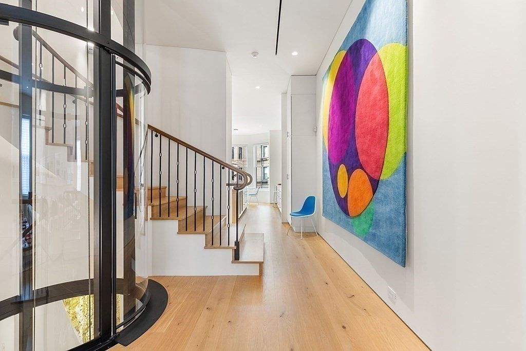 This interior shot showcases a modern hallway with light hardwood flooring and white walls. A unique glass-encased elevator is visible alongside a traditional staircase with dark metal railings and wooden steps. A vibrant, abstract art piece hangs on the wall, adding a pop of color to the space, and a blue chair sits in the hallway.
