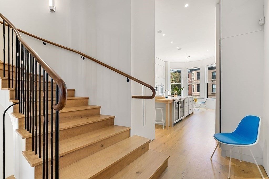 This interior shot showcases a modern hallway with wooden stairs and a black metal railing. The walls are painted white, creating a bright and airy atmosphere. The hallway leads into a kitchen area, offering a glimpse of the home's open floor plan and contemporary design.