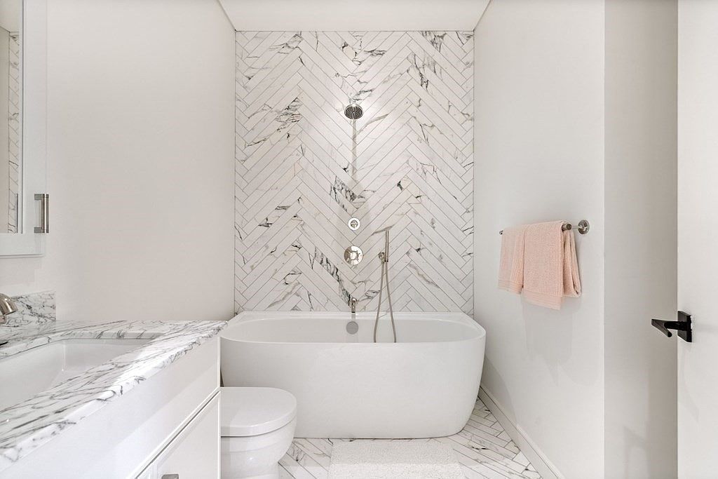 This is a bright and modern primary bathroom featuring a freestanding white bathtub with a marble herringbone tile accent wall. The bathroom also includes a toilet, a sink with marble countertops, and a towel rack with neatly folded towels. The overall aesthetic is clean, luxurious, and spa-like, creating a relaxing atmosphere.