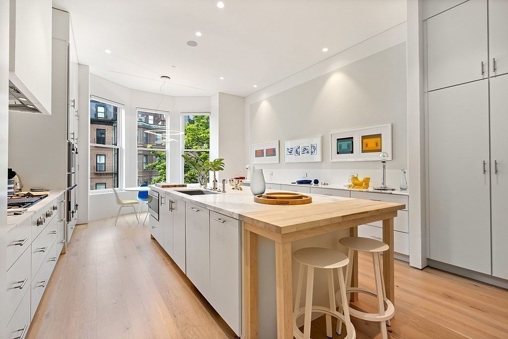 This is a bright, modern kitchen featuring white cabinetry, stainless steel appliances, and a large island with a butcher block extension. The kitchen has hardwood floors and is well-lit with recessed lighting and natural light from the windows. The perspective is from the side, showcasing the length of the kitchen and the island.