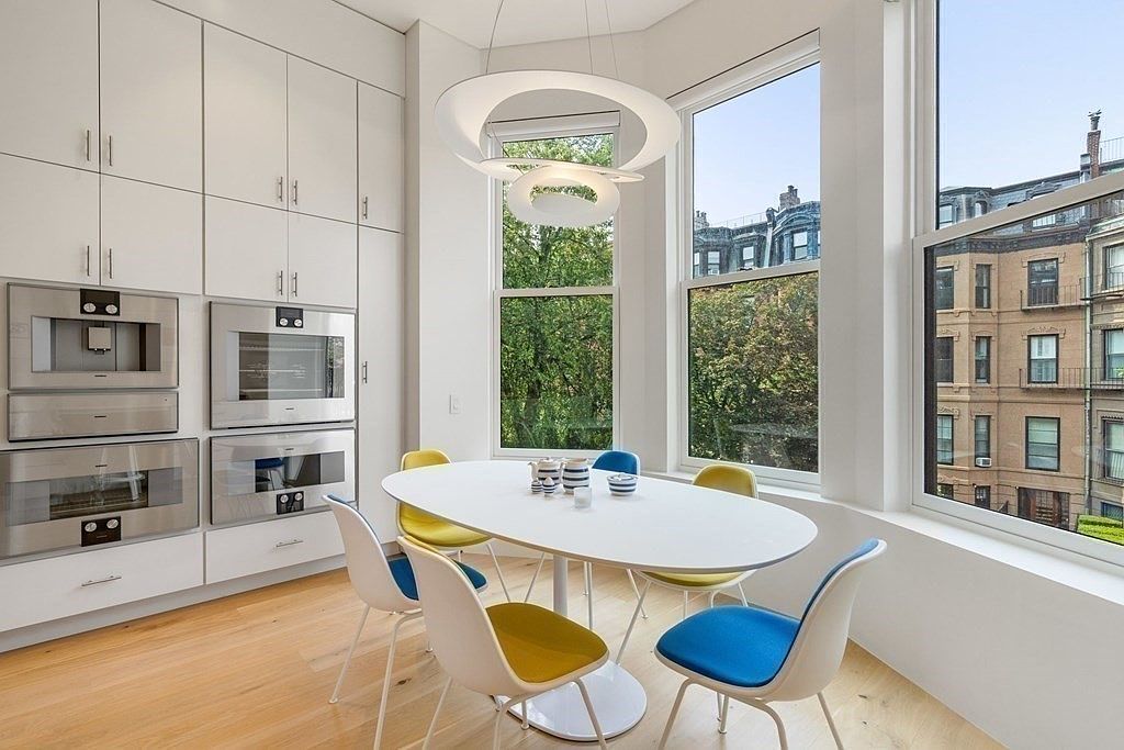 This is a bright and modern dining room featuring a white oval table surrounded by chairs with blue and yellow cushions. Large windows offer a view of the outdoors, and a unique circular chandelier hangs above the table. Built-in white cabinets and stainless steel appliances line one wall, creating a clean and contemporary aesthetic.