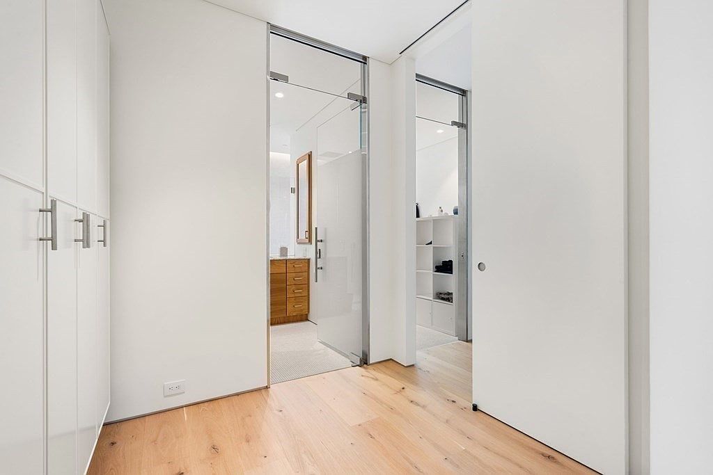 This interior shot showcases a modern hallway with light hardwood flooring and white walls, leading to a bathroom and a walk-in closet. The bathroom is visible through a glass sliding door, revealing a wooden vanity, while the closet is accessible through a white sliding door. The space is clean, bright, and minimalist, emphasizing functionality and contemporary design.