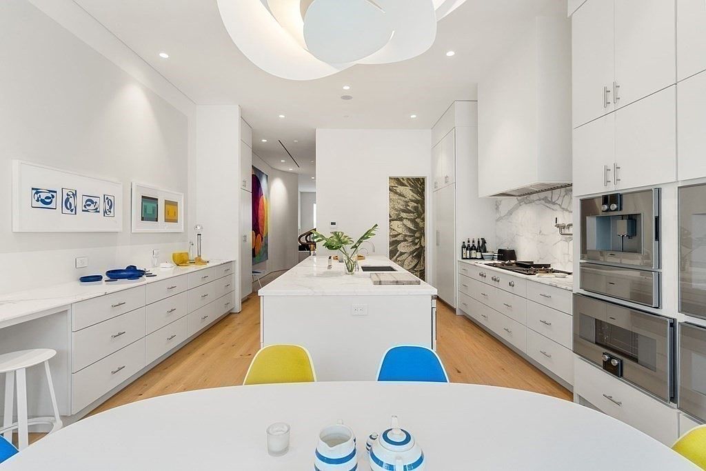 This is a bright, modern kitchen with white cabinetry, stainless steel appliances, and a large central island with a marble countertop. The kitchen features a unique ceiling light fixture and is accented with colorful chairs and artwork. The space is open and airy, creating a clean and inviting atmosphere.
