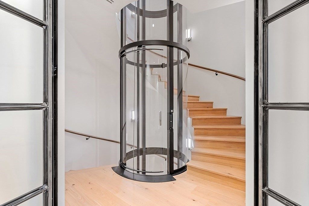 This interior shot showcases a modern hallway with a unique cylindrical glass elevator as a focal point. Adjacent to the elevator, wooden stairs ascend, complemented by a sleek handrail. The space is well-lit and features a clean, minimalist design, creating an impression of contemporary luxury and accessibility.