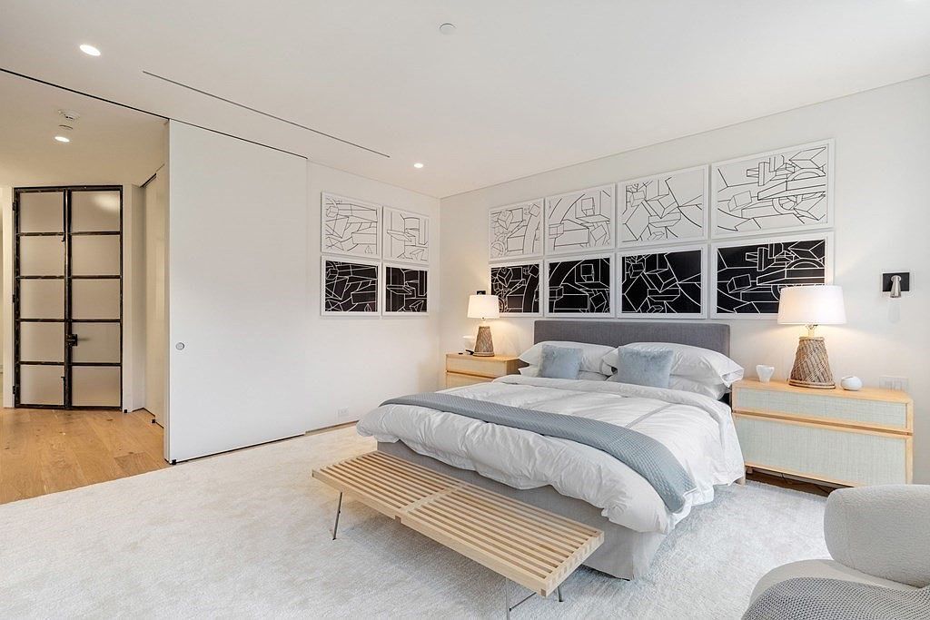 This is a bright and airy primary bedroom featuring a large bed with a gray headboard, white linens, and light blue accent pillows. The room is decorated with a series of black and white abstract art pieces above the bed, and light wood nightstands flank the bed, each with a lamp. A light wood bench sits at the foot of the bed, and the room is carpeted in a light color, creating a serene and inviting atmosphere.