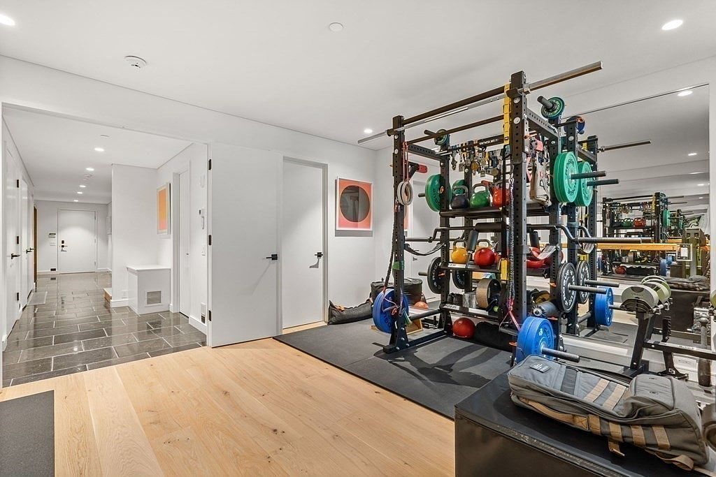 This is an interior shot of a home gym featuring a weight rack with various weights and kettlebells. The room has light wood flooring and a mirrored wall, which visually expands the space. A hallway with dark tile flooring is visible in the background, leading to other rooms.