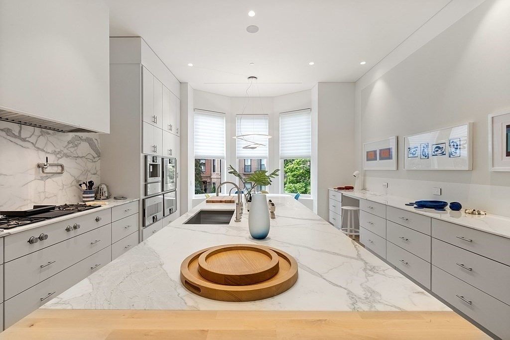This is a bright, modern kitchen featuring a large island with a marble countertop and light wood accents. The kitchen includes stainless steel appliances, white cabinetry, and a marble backsplash. Natural light floods the space through a bay window, creating a clean and inviting atmosphere.