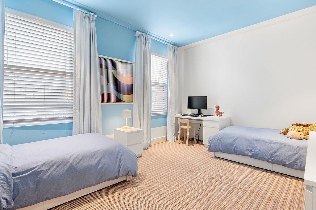 This is a bright and airy guest bedroom featuring two twin beds with blue bedding, a shared desk area with a computer, and a neutral-toned striped rug. The walls are painted in a combination of white and light blue, with white trim and curtains, creating a serene and inviting atmosphere. The room is well-lit by natural light from the windows.