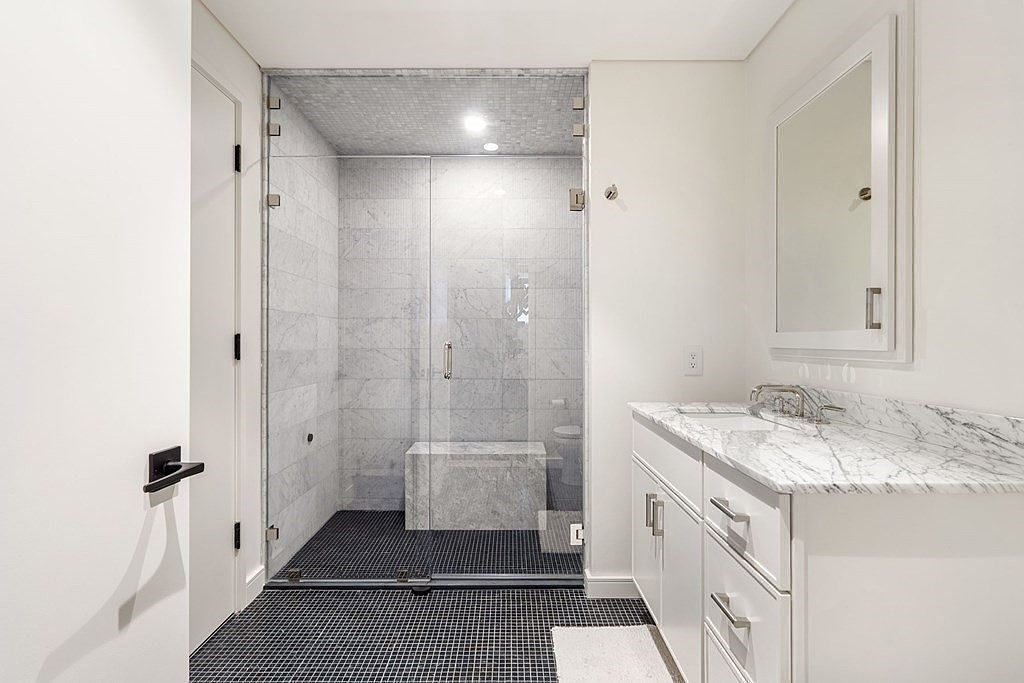 This is a well-lit primary bathroom featuring a glass-enclosed shower with marble tile walls and a built-in bench. The floor is covered in dark mosaic tiles, contrasting with the white walls and vanity. The vanity has a marble countertop and white cabinets with silver hardware, complemented by a framed mirror above.