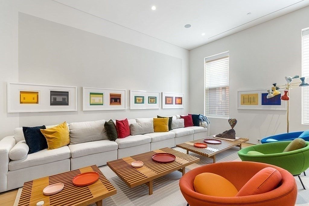 This is a bright and modern living room featuring a large, modular white sofa adorned with colorful pillows. The room is decorated with framed artwork and unique, colorful chairs, creating a vibrant and inviting atmosphere. Several wooden slat coffee tables are arranged in front of the sofa, adding a touch of natural texture to the space.