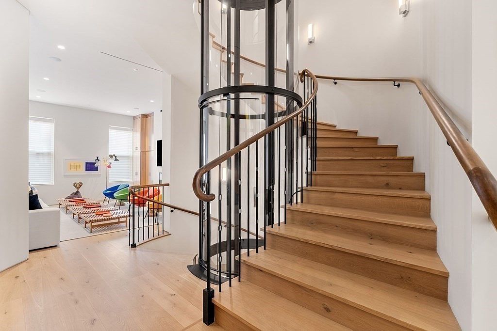 This interior shot showcases a modern staircase with wooden steps and a unique cylindrical glass elevator. The staircase features a dark metal railing with a wooden handrail, complemented by light-colored hardwood flooring. The adjacent living area is visible, adding depth and context to the space.