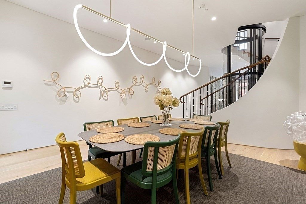 This interior shot showcases a modern dining room with a long table set for a meal, surrounded by stylish chairs in varying shades of green and yellow. A unique, linear light fixture hangs above the table, complemented by an artistic wall decoration. The room features a staircase in the background, adding architectural interest and depth to the space.
