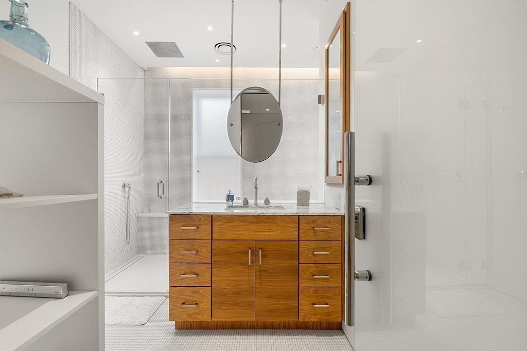This is a well-lit primary bathroom featuring a wooden vanity with a marble countertop and an oval mirror suspended from the ceiling. The bathroom includes a glass-enclosed shower with white tiled walls and a modern design. The overall impression is clean, modern, and luxurious.
