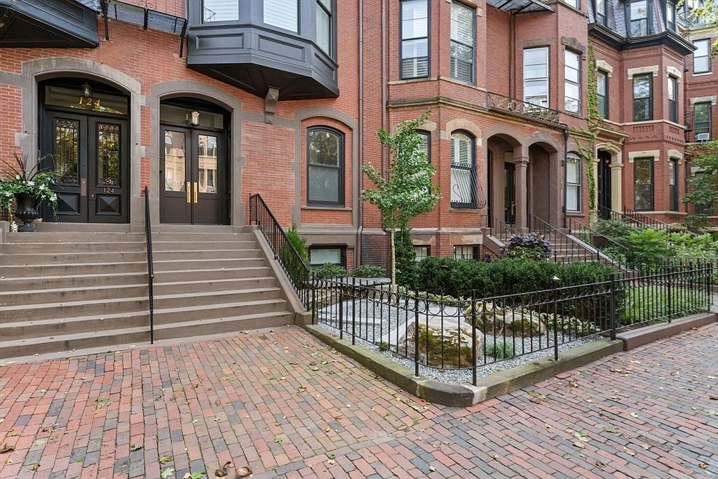 This image showcases the front exterior of a classic brick townhouse. The building features arched doorways, detailed window frames, and a well-maintained brick walkway leading to the entrance. The landscaping includes a small garden area enclosed by a wrought iron fence, adding to the property's curb appeal and charm.