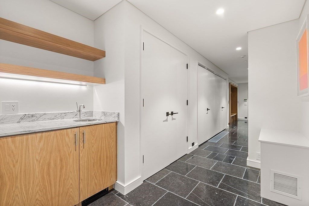 This interior shot showcases a modern hallway with dark gray tiled flooring and white walls. A built-in wet bar with light wood cabinetry and marble countertops is visible on the left, while several white doors line the hallway, leading to other rooms. The space is well-lit with recessed lighting, creating a clean and sophisticated atmosphere.