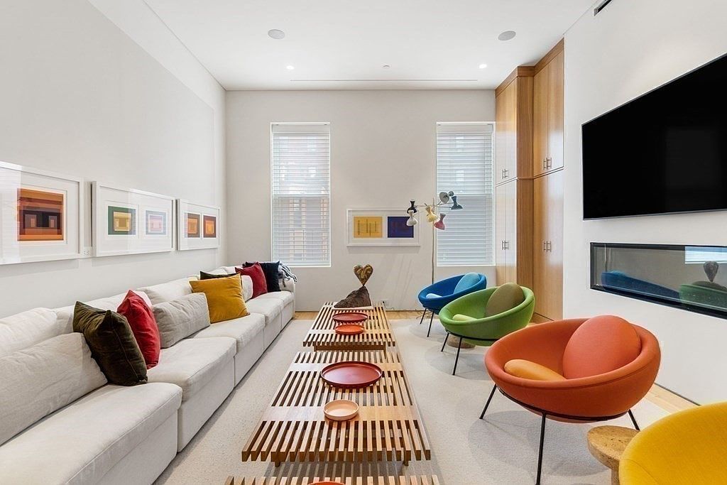 This is an interior shot of a modern living room featuring a long white sectional sofa adorned with colorful pillows, complemented by a series of framed artwork on the wall. A unique slatted coffee table runs the length of the sofa, decorated with decorative plates, while colorful pod chairs add a playful touch to the space. The room is well-lit with natural light from the windows and features a modern fireplace and TV.