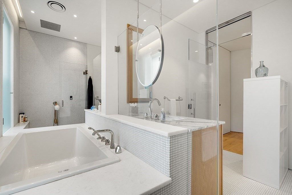 This is a bright and modern primary bathroom featuring a large soaking tub, a vanity with a marble countertop and mosaic tile backsplash, and a glass-enclosed shower. The color palette is primarily white, creating a clean and spa-like atmosphere. A round mirror hangs above the vanity, adding a touch of elegance.