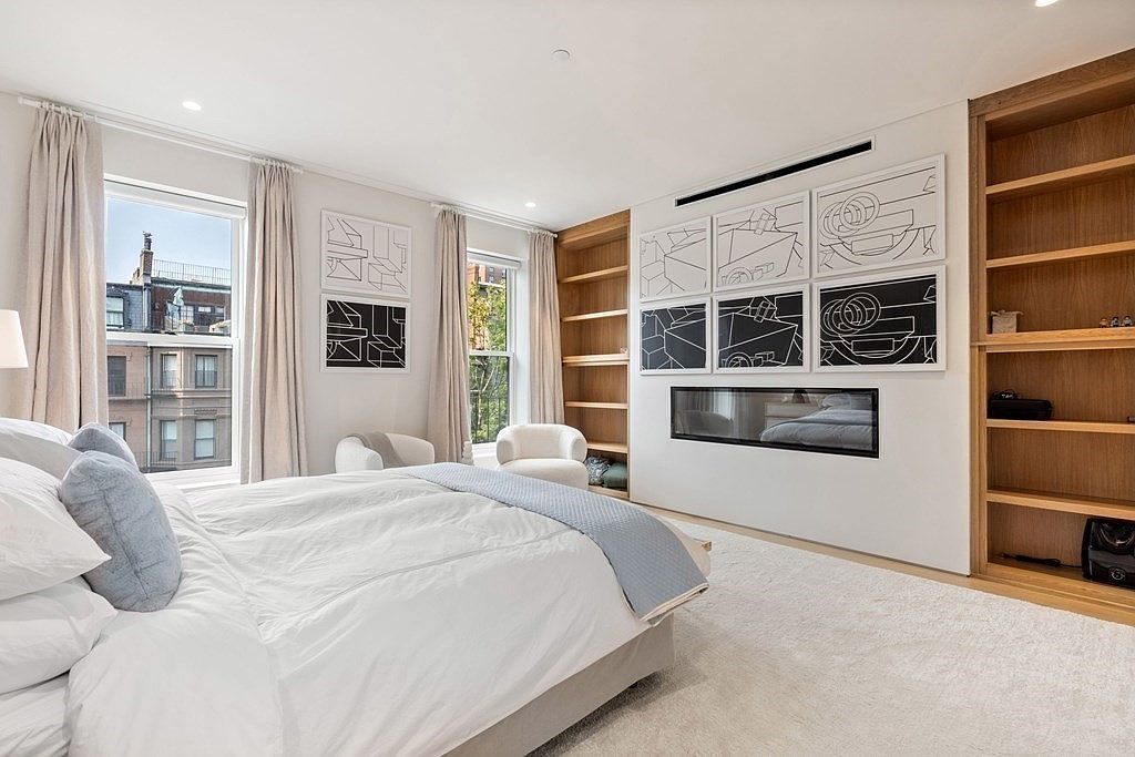 This is a bright and airy primary bedroom featuring a large bed with white linens and blue accent pillows. The room has two windows with beige curtains, built-in wooden bookshelves, and modern black and white artwork above a sleek fireplace. The overall impression is clean, modern, and inviting.