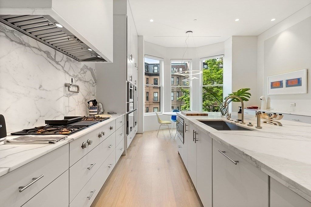 This is a bright, modern kitchen featuring white cabinetry and marble countertops. Stainless steel appliances and a gas range are visible along one wall, while a large sink and faucet are featured on the island. A bay window provides natural light and a view of the exterior, enhancing the kitchen's open and airy feel.