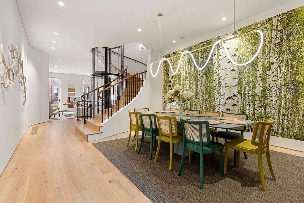 This interior shot showcases a modern dining room with a unique blend of natural and contemporary elements. The room features a dining table surrounded by stylish, two-toned chairs, complemented by a striking light fixture overhead. A birch tree wallpaper adds a touch of nature, while a curved staircase with a glass elevator creates a sophisticated architectural focal point.