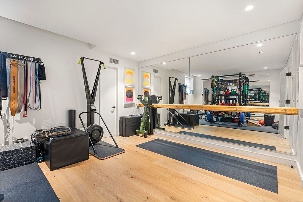 This is a well-equipped home gym featuring a variety of exercise equipment, including a ski erg, weight rack, and free weights. A large mirror covers one wall, enhancing the sense of space and allowing for form checks during workouts. The room has light wood flooring and white walls, creating a bright and functional workout environment.