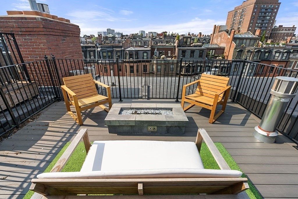 This image showcases a stylish rooftop deck or balcony, furnished with two wooden chairs, a modern fire pit, and a cushioned bench. The deck features a dark-toned wooden surface and a black metal railing, offering views of the surrounding cityscape. The overall impression is one of urban sophistication and relaxation.