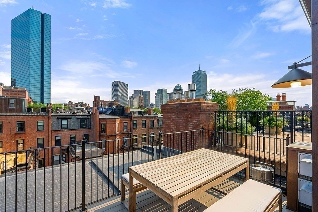 This image showcases a rooftop deck or balcony with a wooden dining table and bench seating, offering an outdoor living space with a cityscape view. The deck features black metal railings and potted plants, adding to the ambiance. The backdrop includes several buildings, including a tall skyscraper, suggesting an urban setting.