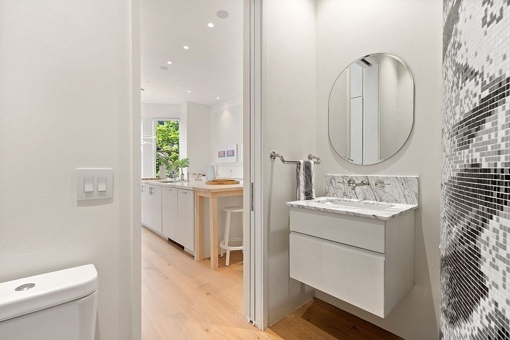 This is a modern guest bathroom featuring a floating vanity with a marble countertop and an oval mirror. The walls are painted in a neutral tone, and there is a mosaic tile accent wall. The bathroom has a clean and minimalist design, with light wood flooring extending into an adjacent room.