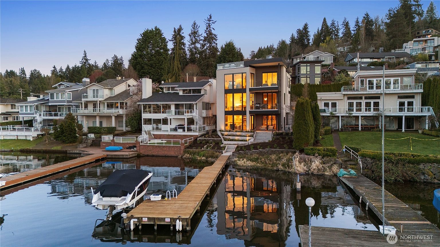 This waterfront property features a modern, multi-story home with expansive glass walls that offer panoramic views of the lake. The exterior showcases a contemporary design with clean lines, a private dock with a boat lift, and tiered landscaping leading down to the water's edge. The warm interior lighting creates an inviting atmosphere, highlighting the home's architectural prominence against the backdrop of a forested hillside.