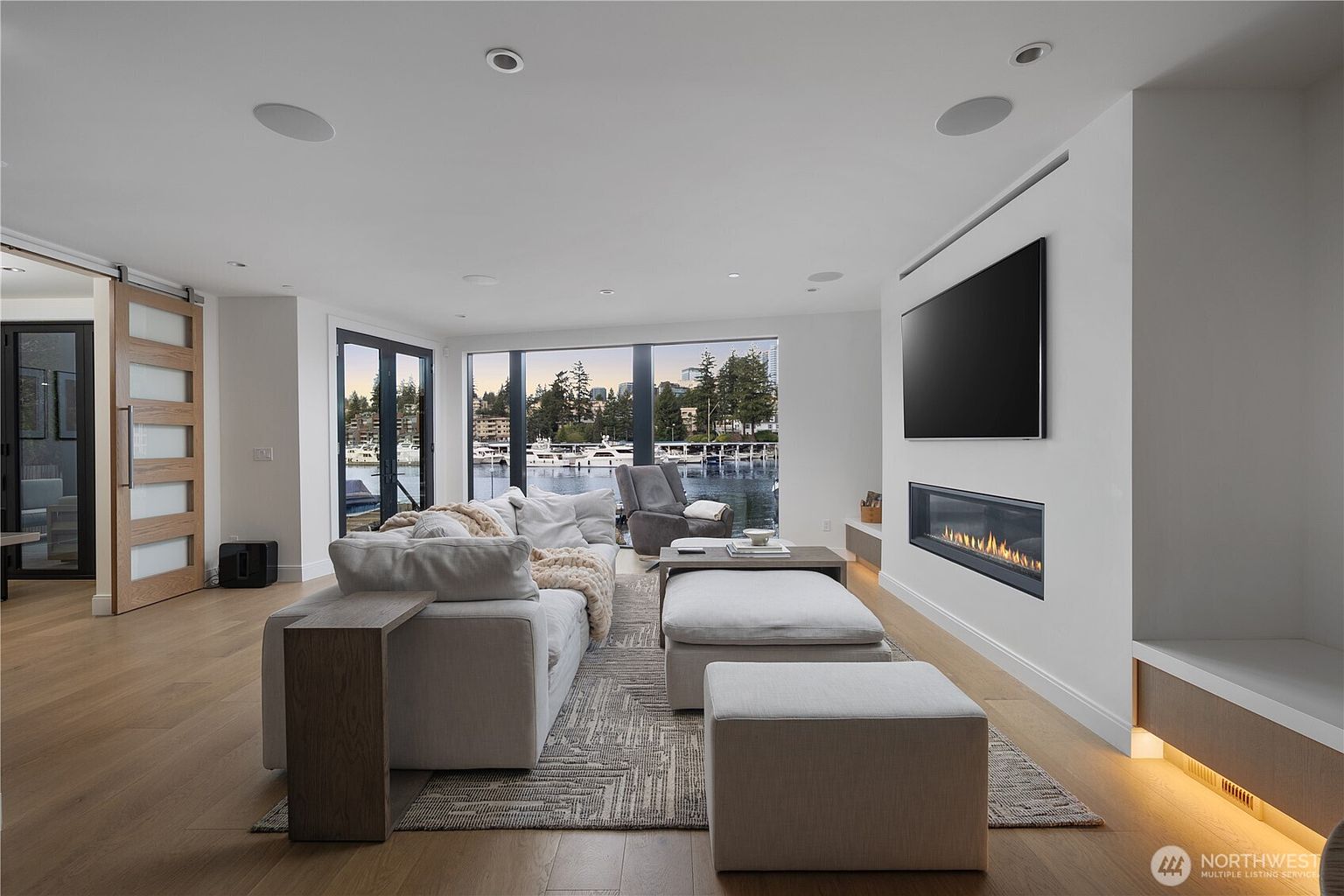 This contemporary living room features a neutral color palette, light wood flooring, and a large sectional sofa arranged around a patterned area rug. A linear fireplace is integrated into the wall beneath a wall-mounted television, while expansive windows provide a scenic view of the water and boats outside. The space is illuminated by recessed lighting and offers a serene, open-concept atmosphere.
