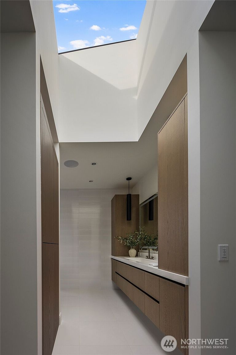 This modern bathroom features a sleek, floating vanity with light wood cabinetry and a clean white countertop. A large skylight above provides abundant natural light, highlighting the minimalist aesthetic and the tall, light wood cabinetry flanking the vanity. The perspective looks down the hallway toward the vanity, emphasizing the clean lines and bright, airy atmosphere of the space.