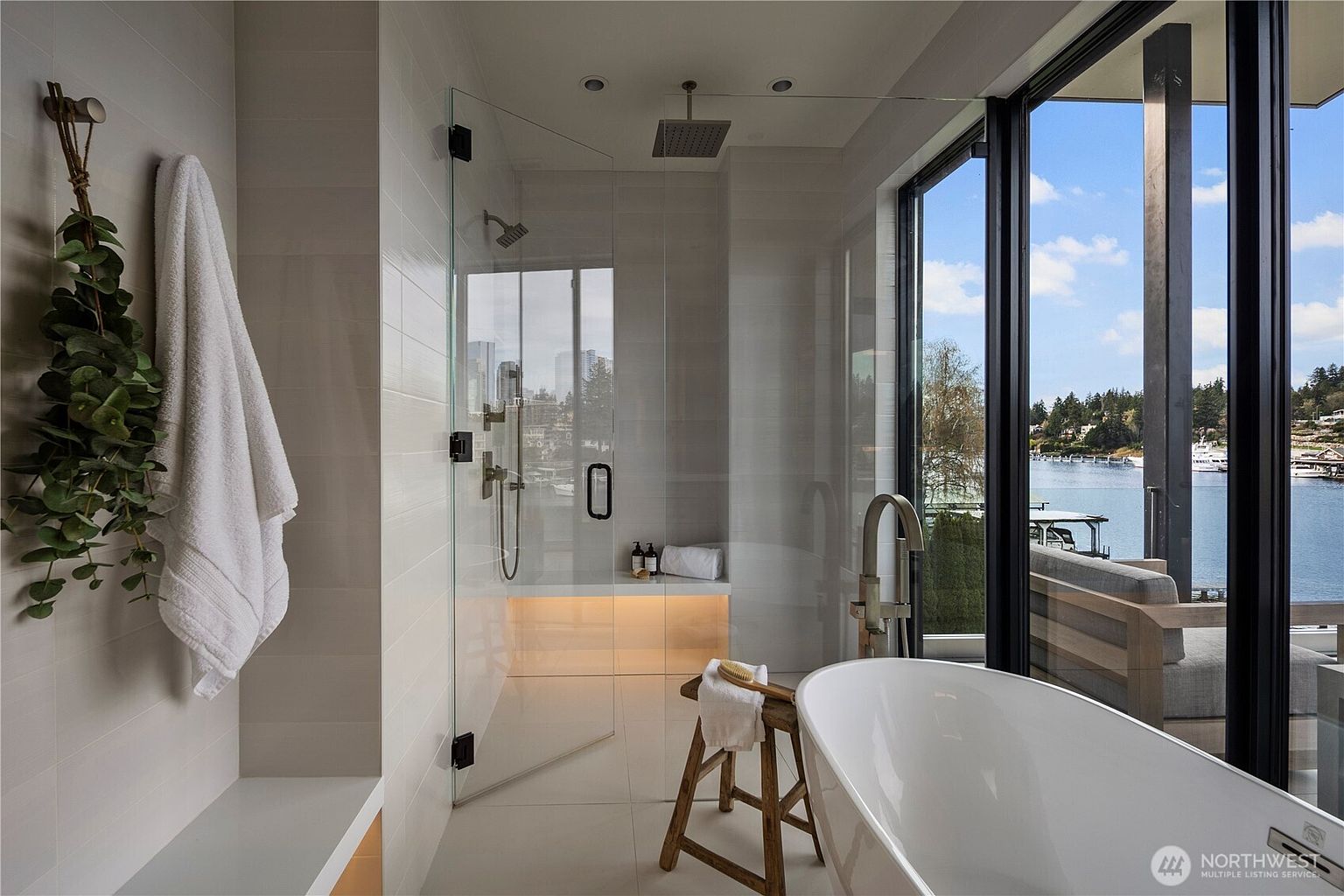 This luxurious primary bathroom features a sleek, glass-enclosed walk-in shower with a built-in bench and integrated lighting, adjacent to a freestanding soaking tub. Large windows provide an expansive, unobstructed view of the water and surrounding landscape, creating a serene and spa-like atmosphere. A decorative wooden stool and a hanging plant add organic textures to the clean, modern aesthetic of the space.