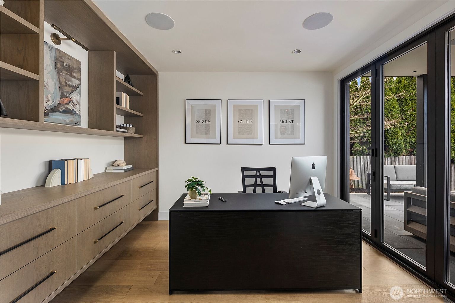 This modern home office features a sleek, dark-toned desk positioned centrally in the room, complemented by custom light wood built-in cabinetry and shelving along the left wall. The space is brightened by large glass doors that provide a seamless transition to an outdoor patio area. Three framed prints on the wall add a sophisticated touch, creating a professional yet inviting workspace.