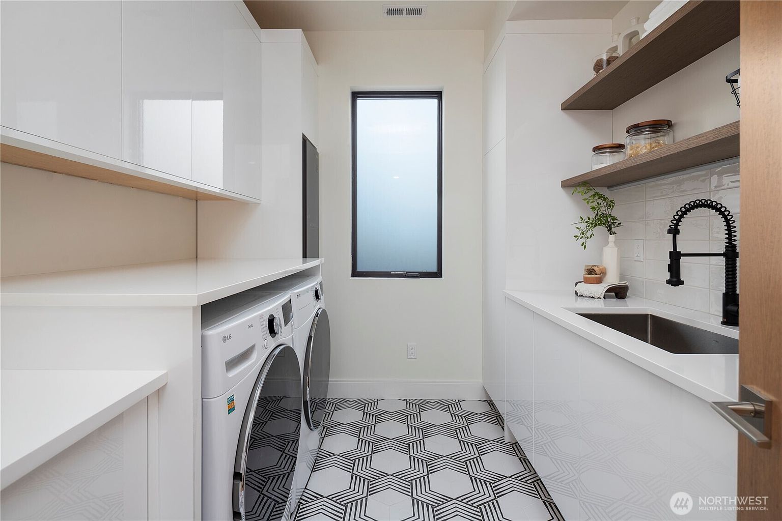 This modern laundry room features sleek white cabinetry, a white countertop, and a built-in sink with a black gooseneck faucet. The space is equipped with a side-by-side washer and dryer set, complemented by open wood shelving and striking geometric patterned floor tiles. A central window with frosted glass provides natural light, creating a clean and functional aesthetic.