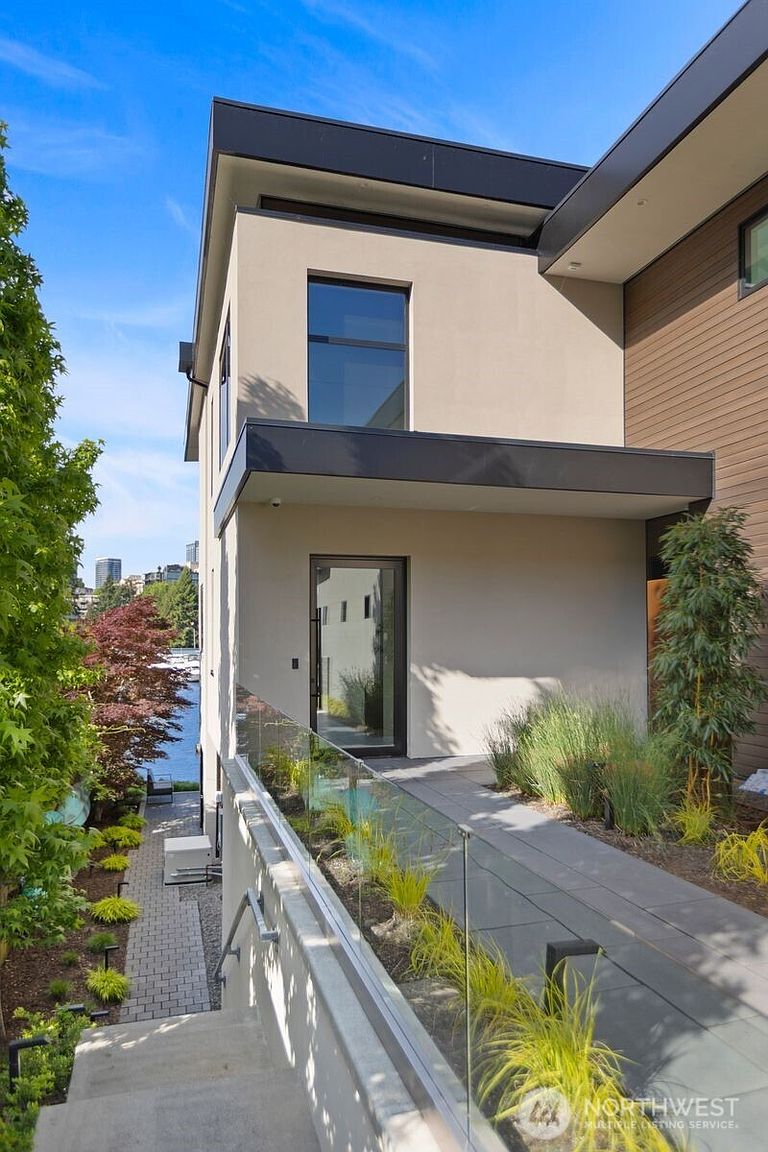 This modern entryway features a sleek glass-paneled door set against a smooth, light-colored facade with dark trim accents. A paved walkway leads to the entrance, bordered by a glass railing and landscaped garden beds with ornamental grasses. The perspective captures the transition from the outdoor stairs to the home's entrance, with a glimpse of a waterfront setting in the background.