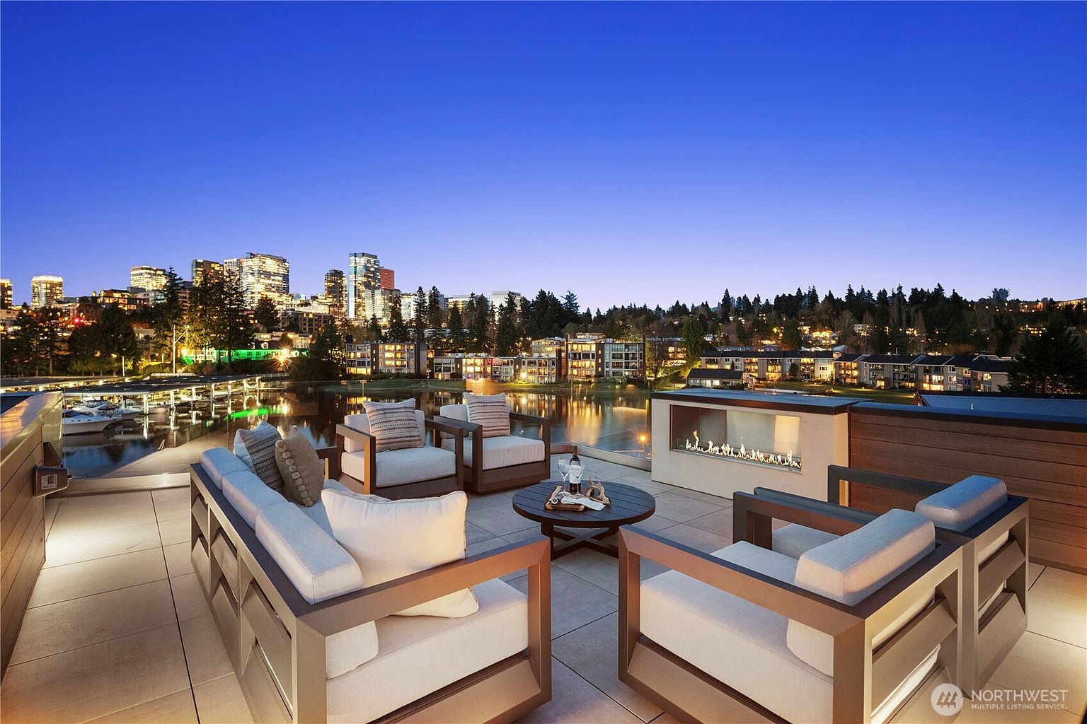This expansive rooftop terrace offers a sophisticated outdoor living space furnished with modern, clean-lined seating arranged around a central circular coffee table. A linear, glass-enclosed gas fireplace provides a focal point, while the elevated vantage point captures a stunning panoramic view of the city skyline and the tranquil water below at dusk. The setting creates an elegant, high-end atmosphere perfect for evening entertaining.
