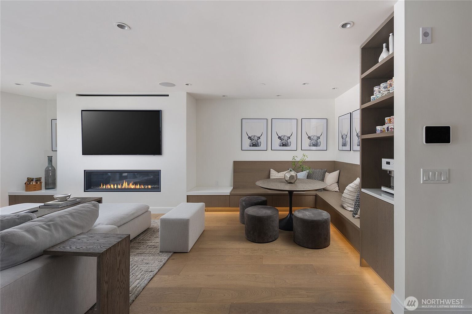 This contemporary living space features an open-concept layout with light wood flooring and a neutral color palette. A linear fireplace is integrated into the wall beneath a wall-mounted television, while an adjacent dining nook offers built-in bench seating and a round table. The room is accented by framed animal prints and a tall shelving unit, creating a cohesive and inviting atmosphere.