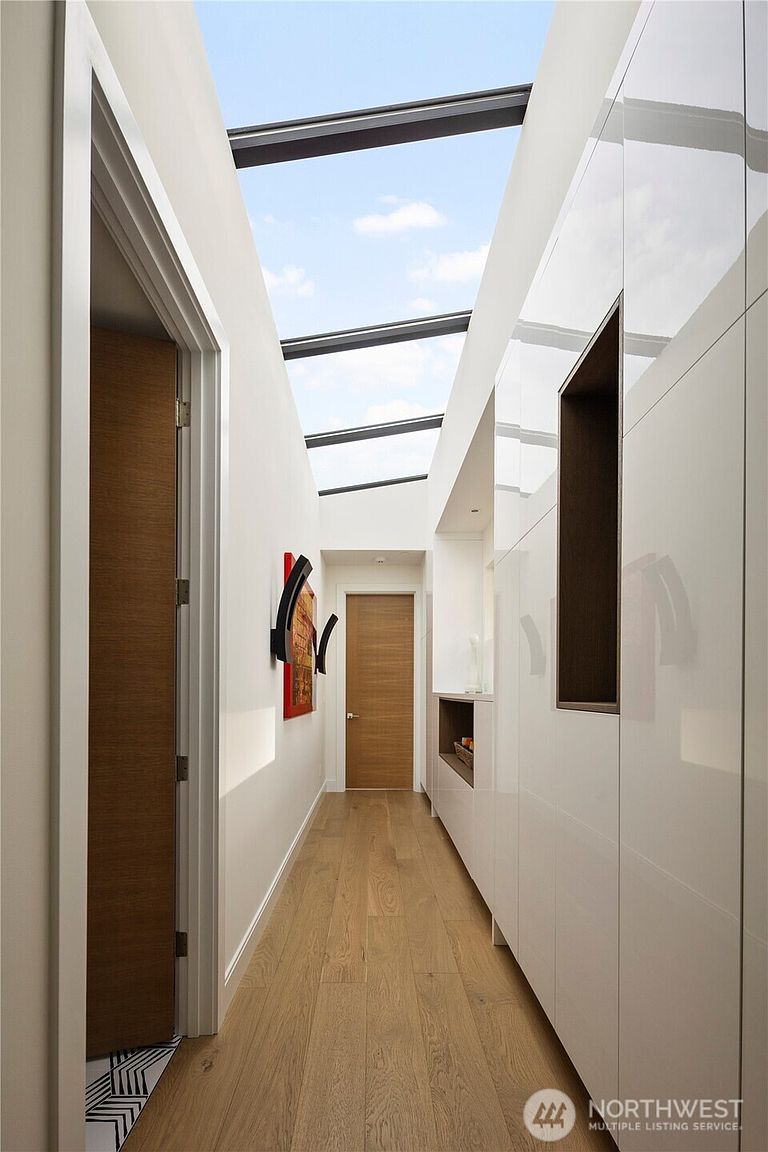 This modern hallway features a striking skylight that runs the length of the ceiling, flooding the space with natural light. The design includes light wood flooring, sleek white cabinetry with integrated shelving, and a warm wood door at the end of the corridor. The minimalist aesthetic is complemented by a piece of abstract wall art, creating a bright and sophisticated transition space.
