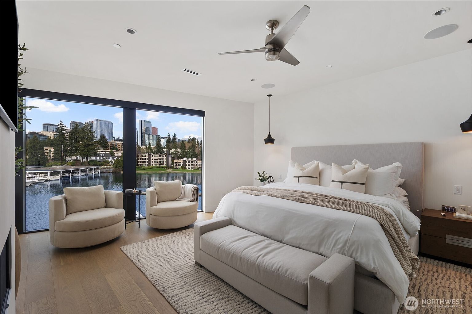 This primary bedroom features a contemporary design with a large bed centered against a neutral-toned wall, complemented by two swivel armchairs positioned near expansive floor-to-ceiling windows. The room offers a serene view of the water and city skyline, enhanced by light wood flooring and a textured area rug. A minimalist ceiling fan and pendant lighting add to the sophisticated, airy atmosphere of the space.