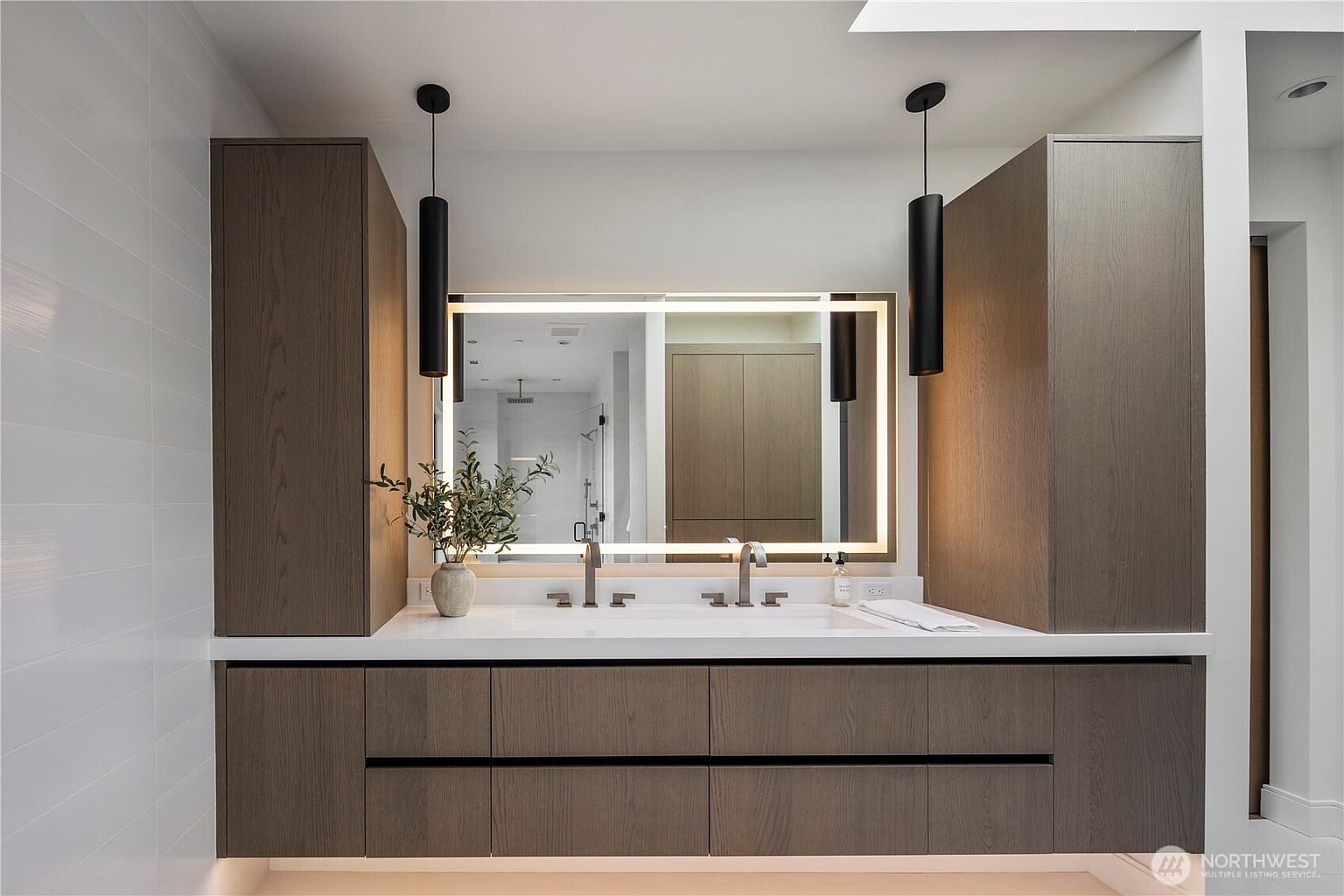 This modern primary bathroom features a double vanity with dark wood cabinetry and a clean white countertop. A large, rectangular LED-backlit mirror is centered between two tall, dark wood storage towers, complemented by sleek black pendant lights. The minimalist design is enhanced by wall-mounted faucets and a floating vanity aesthetic, creating a sophisticated and contemporary atmosphere.