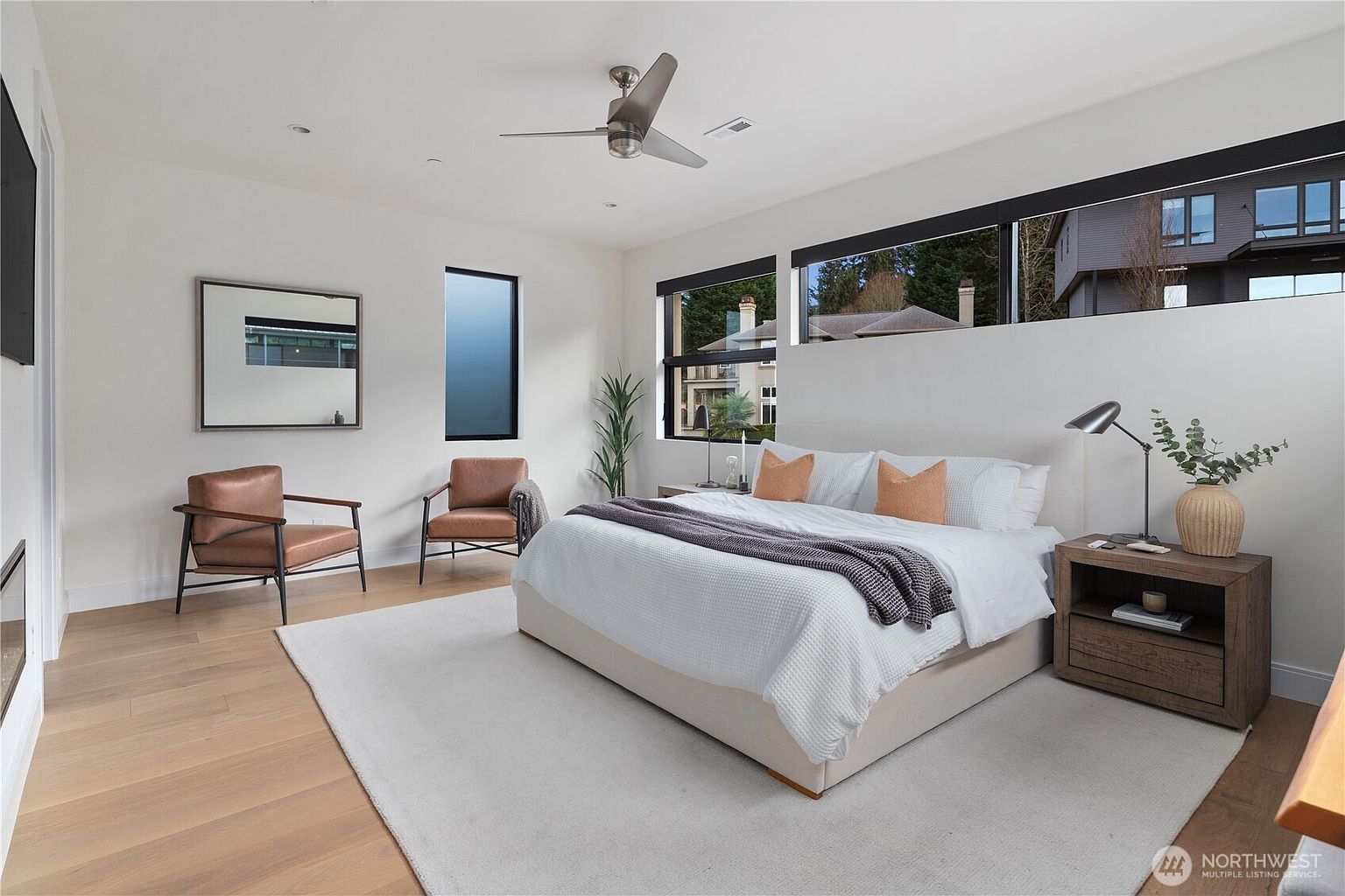 This spacious primary bedroom features a modern aesthetic with light wood flooring, white walls, and a large area rug. The room is furnished with a king-sized bed, two leather armchairs, and a wooden nightstand, all illuminated by a ceiling fan and a sleek bedside lamp. Large horizontal windows provide natural light and views of the surrounding neighborhood, creating a bright and airy atmosphere.