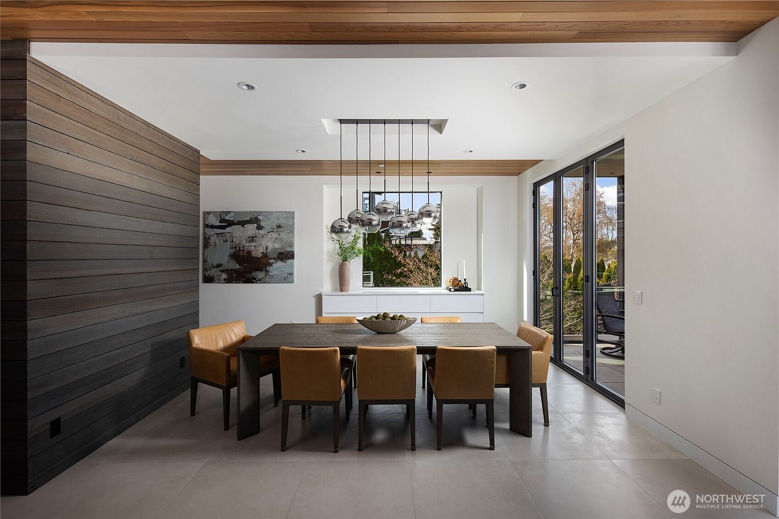 This contemporary dining room features a large, dark wood table surrounded by eight leather-upholstered chairs, set against a striking accent wall of horizontal wood planks. A modern multi-globe pendant light fixture hangs above the table, while a white sideboard sits beneath a large window that provides natural light and a view of the outdoors. The space is completed by large glass doors that open to an adjacent patio, creating a seamless transition between indoor and outdoor living.