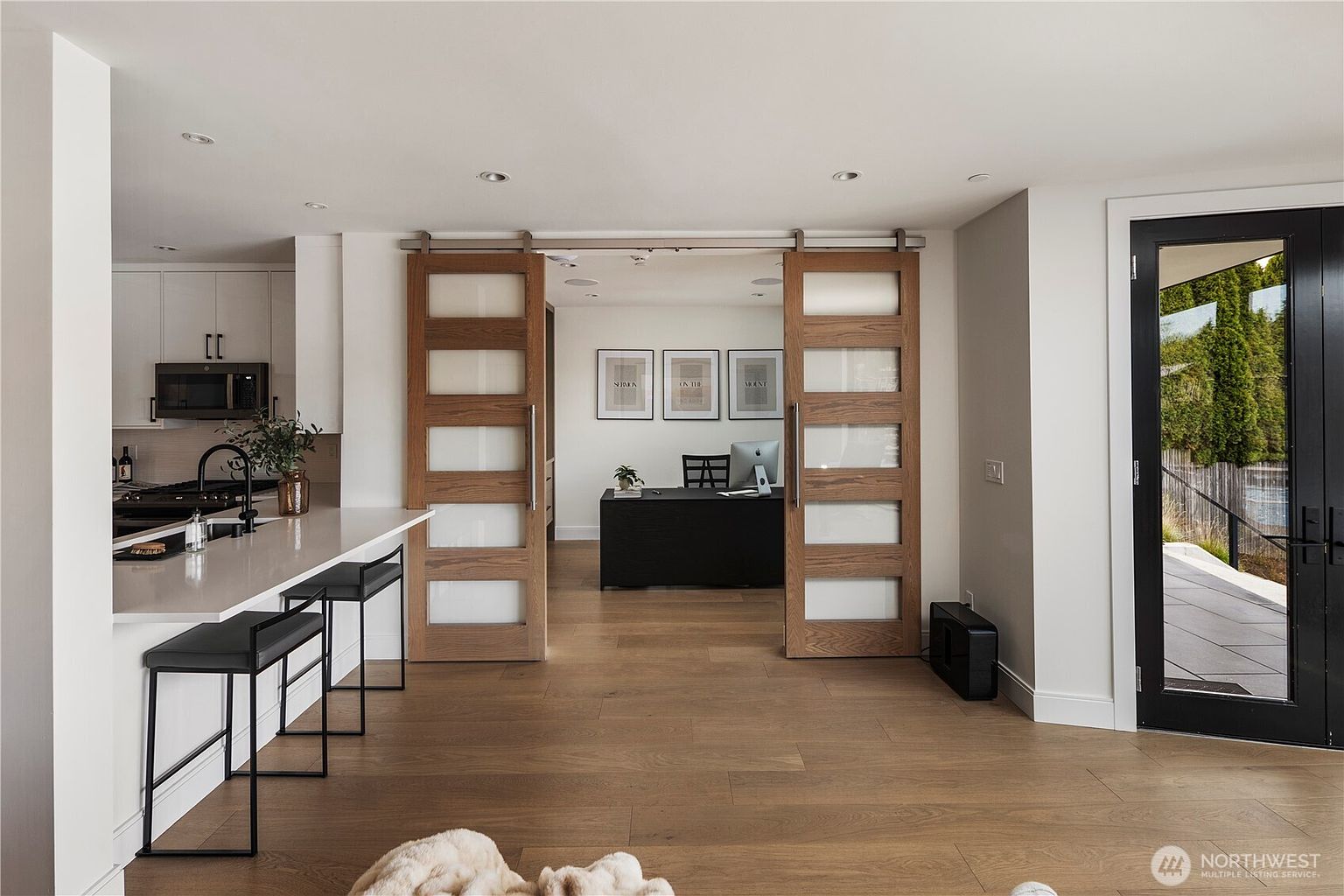 This open-concept space features a modern kitchen with white cabinetry and a breakfast bar, seamlessly transitioning into a home office through dual sliding wood-framed glass doors. The room is anchored by light-toned wood flooring and a neutral color palette, creating a bright and airy atmosphere. A black exterior door on the right provides a sharp contrast, while the office beyond is furnished with a dark desk and minimalist wall art.