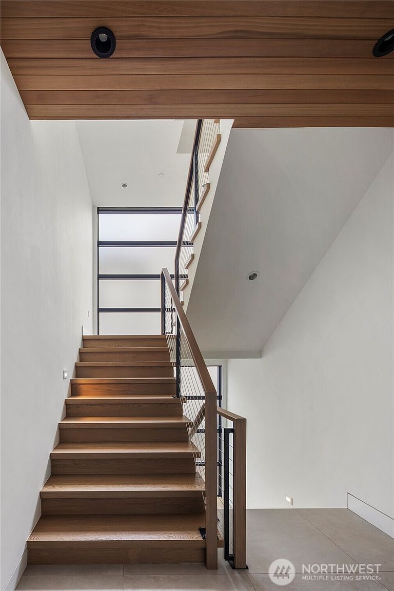 This modern staircase features light wood treads and a sleek railing system with thin vertical cables, creating an open and airy aesthetic. The space is defined by crisp white walls and a warm wood-paneled ceiling, with a large window at the landing providing natural light. The perspective is from the base of the stairs, looking upward to highlight the clean lines and architectural transition between levels.