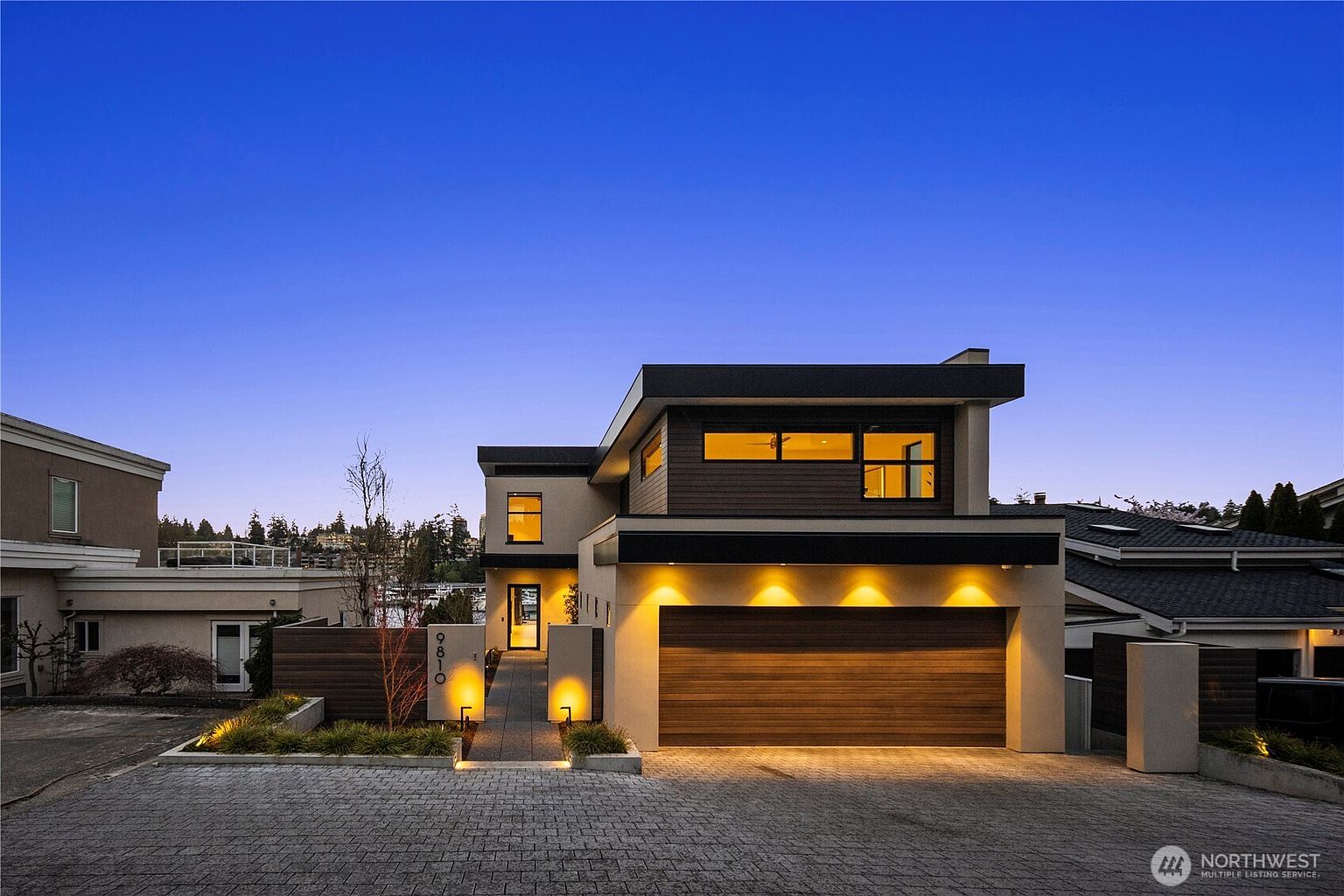 This modern two-story residence features a striking contemporary design with a mix of smooth stucco and horizontal wood siding. The front facade is highlighted by warm, recessed exterior lighting that accentuates the garage door and the entryway path. A paved driveway leads to the garage, while the clean lines and geometric architecture create a sophisticated and inviting curb appeal at dusk.