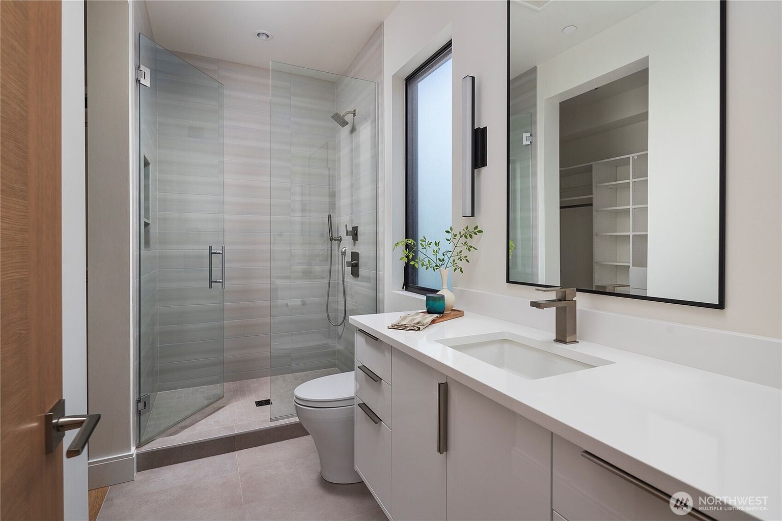 This modern bathroom features a sleek vanity with a white countertop, a rectangular undermount sink, and a tall, thin mirror with a black frame. To the left, a glass-enclosed walk-in shower showcases light-toned horizontal tile work and a handheld showerhead. The space is bright and airy, complemented by a tall window that allows natural light to filter into the room.