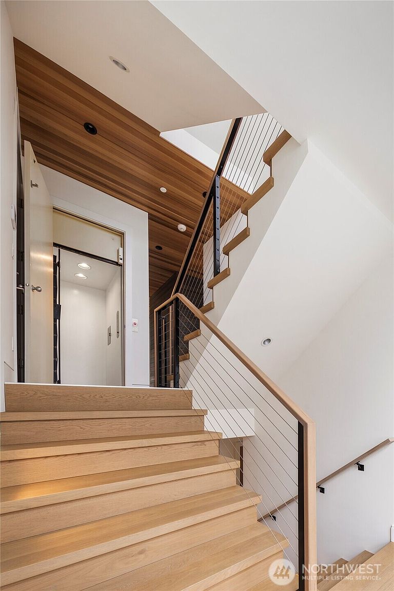 This modern staircase features light wood treads and a sleek railing system with thin metal balusters. The ceiling above the stairs is finished with warm-toned wood planks, adding a natural texture to the bright, white-walled space. A doorway to the left reveals an elevator, highlighting the home's accessibility and contemporary design.