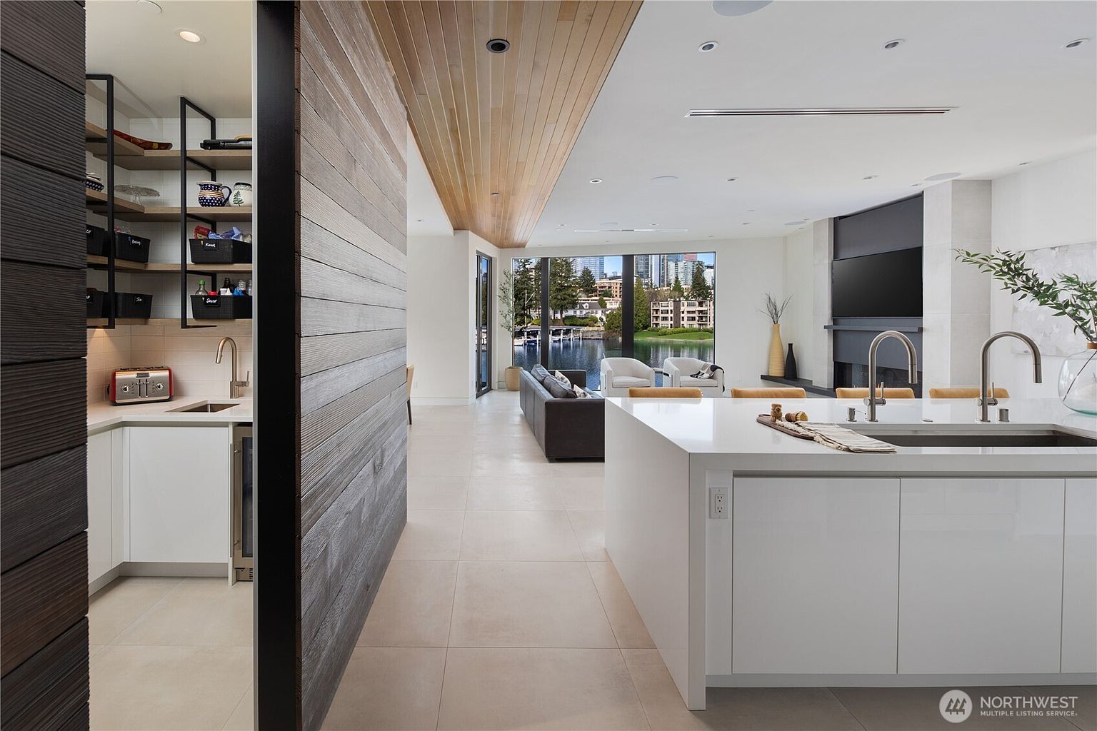 This modern, open-concept kitchen features a large white island with a double sink and a sleek, minimalist aesthetic. To the left, a wood-paneled wall separates the main kitchen area from a walk-in pantry with open shelving and a small prep sink. The space flows seamlessly into a living area with large windows that provide a clear view of the water and surrounding landscape.