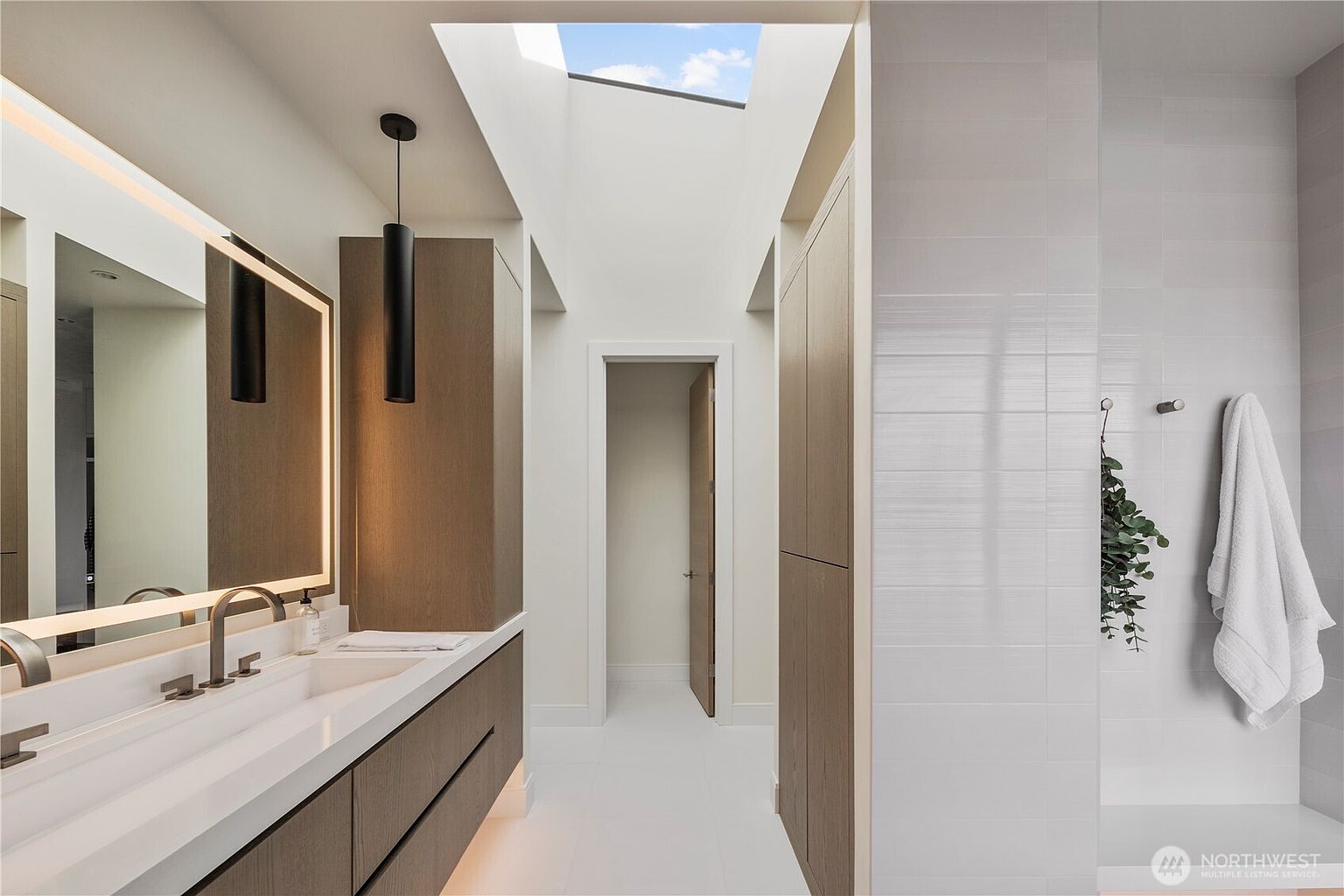 This modern primary bathroom features a sleek vanity with a light wood finish, a large illuminated mirror, and a minimalist faucet. A skylight above the central walkway floods the space with natural light, while a tiled shower area to the right includes a wall-mounted towel hook and decorative greenery. The clean lines and neutral color palette create a bright, sophisticated, and airy atmosphere.